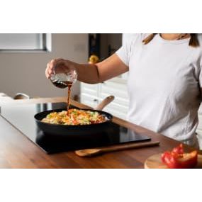 Action shot of using the Pyrex Cook & Care recycled aluminium frying pan to cook, capturing a user pouring sauce over vegetables, great for healthy cooking.